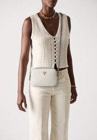 Geantă crossbody albă, texturată, cu un logo triangular, curea din lanț argintiu și închidere cu fermoar, purtată cu un vest tricotat și pantaloni crem.