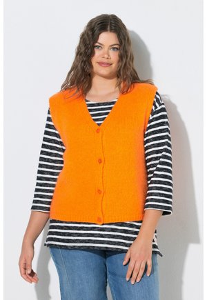 Frau mit welligem braunem Haar, die eine orangefarbene, geknöpfte Weste über einem schwarz-weiß gestreiften Langarmshirt und blauen Jeans vor weißem Hintergrund trägt.
