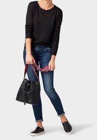 Person, die ein schwarzes langärmliges Oberteil, hochgekrempelte blaue Jeans und schwarze Slipper trägt, hält eine schwarze Bucket Bag mit einem bunten Gurt.