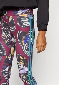 Leggings avec une base bordeaux, ornés d'un motif de papillon dans différentes couleurs, accentués de bandes latérales teal et d'un tissu lisse et extensible.