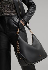 Borsa a spalla in pelle nera con finitura testurizzata, dettagli in oro e logo embossato "JOOP!" sulla tracolla. Forma moderna e curva.