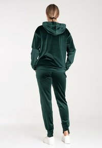 Samtsch trackuit in Dunkelgrün, bestehend aus einem Kapuzenoberteil und taillierten Hosen, mit einer glatten Textur und kontrastierenden Manschetten an den Knöcheln.