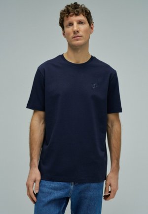 Hombre con cabello rizado vistiendo una camiseta azul marino lisa y jeans azules contra un fondo gris neutro.