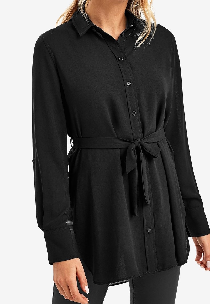 Femme portant un chemisier noir à manches longues boutonné avec col et ceinture nouée à la taille, associé à un pantalon noir, sur fond blanc.