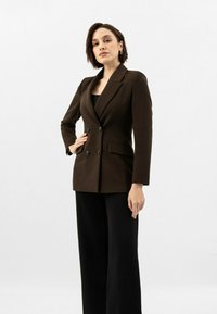 Brązowa marynarka dwurzędowa z klapami, kieszeniami bocznymi i czarnymi guzikami, w połączeniu z czarnymi spodniami o szerokich nogawkach. Gładka tekstura materiału.