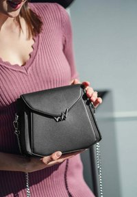 Černá kožená crossbody taška s klopou, s kovovým logem a stříbrným řetízkem, kterou drží osoba v růžovém topu.