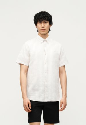 Młody mężczyzna stoi, ubrany w jasną, w paski koszulę na krótki rękaw z guzikami oraz czarne krótkie spodenki na jednolitym jasnym tle.