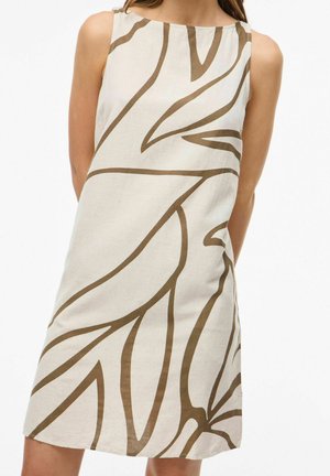 Bezrukávové béžové šaty so širokými hnedými abstraktnými vzormi listov, ktoré nosí osoba stojaca s rukami za chrbtom.