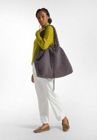 Grande borsa a tracolla grigia in peluche con chiusura a coulisse e superficie texturizzata, abbinata a pantaloni chiari e a un maglione verde a maniche lunghe.