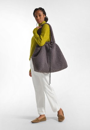 Grand sac fourre-tout en peluche gris avec fermeture à cordon et surface texturée, associé à un pantalon clair et un pullover vert à manches longues.
