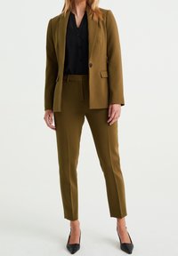 Olivově zelený oblek na míru s úzkým střihem, se sakem na jedno tlačítko a zúženými kalhotami. Doplněn černou košilí na knoflíky a lodičkami.