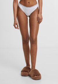 Witte katoenen slip met een kanten rand op de tailleband, gecombineerd met bruine pluizige slippers. Eenvoudig ontwerp, zachte textuur.