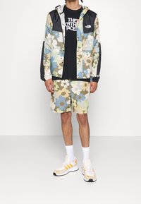 Kurtka i szorty w kwiatowy wzór w pastelowych kolorach; czarne elementy na kurtce; zestawione z białymi skarpetkami i beżowymi trampkami z pomarańczowymi akcentami.