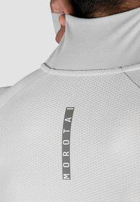 Hellgraues Hemd mit hohem Kragen aus strukturiertem Material, das auf dem Rücken ein vertikales graues Logo-Label "MOROTAI" trägt.