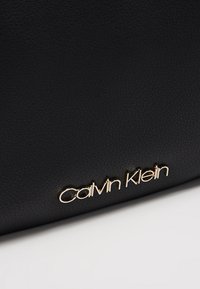 Czarna skórzana torebka z teksturowanym wykończeniem, z wyraźnie widocznym metalicznym złotym logo "Calvin Klein".