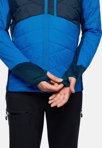 Veste isolante bleue avec surpiqûres en zigzag, accents bleu foncé, fermeture éclair et pouces à l'ouverture des poignets, portée sur un pantalon noir avec poches.