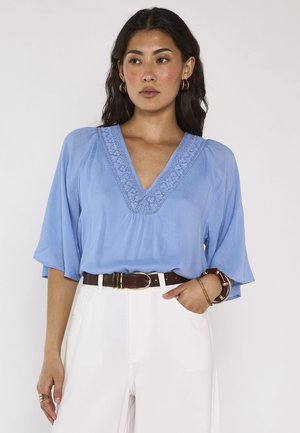 Donna con lunghi capelli scuri che indossa una camicetta azzurro chiaro con scollo a V ricamato, pantaloni bianchi, cintura scura e braccialetti, in piedi contro uno sfondo semplice.