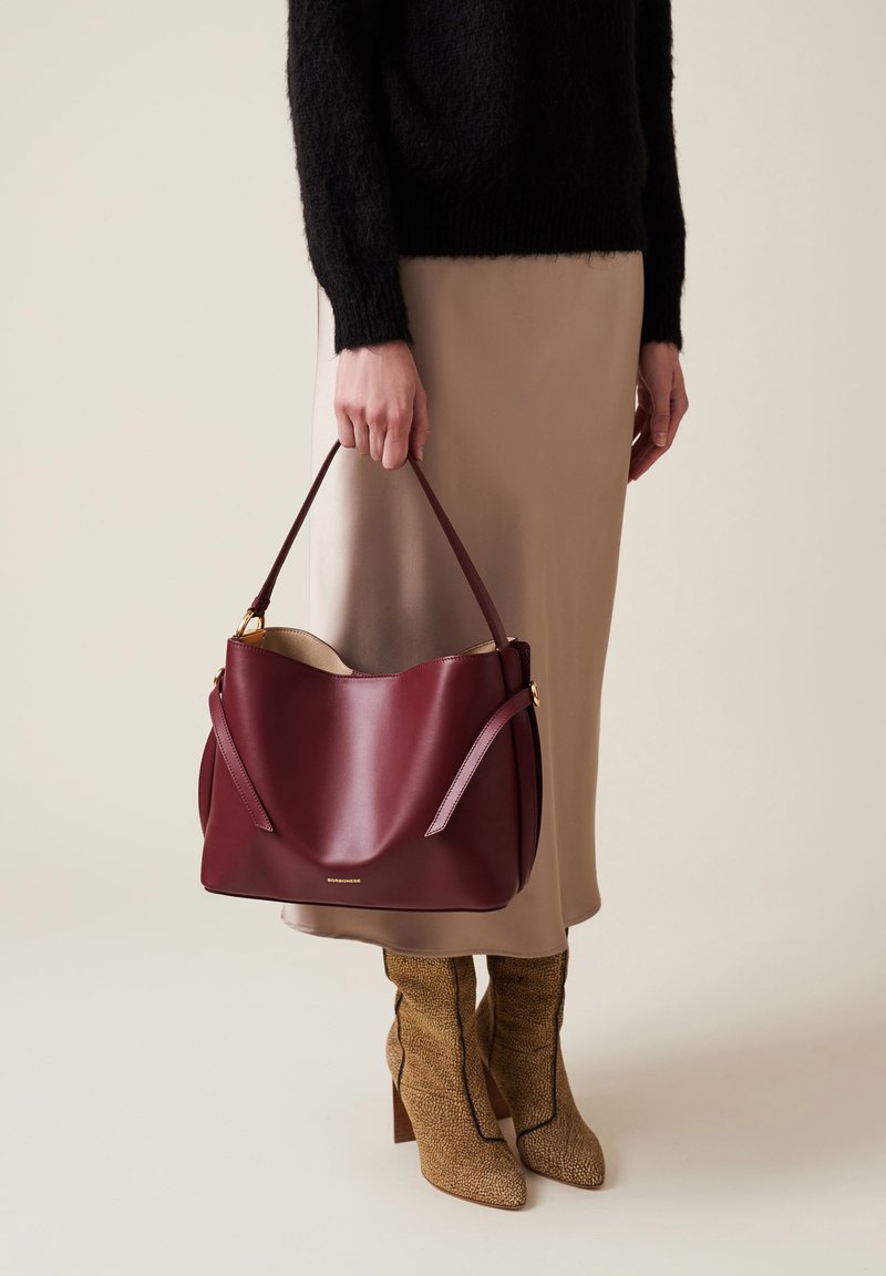 Sac en cuir bordeaux avec une finition lisse, doté d'une double poignée et de quincaillerie dorée. Porté avec une jupe beige et des bottes à motifs.