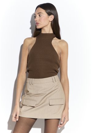 Mujer con un top marrón sin mangas con textura acanalada y minifalda beige con bolsillos con solapa, de pie frente a un fondo blanco, mirando hacia un lado.