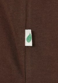Brauner Baumwollstoff mit einem weißen Etikett, das ein grünes Blattemuster zeigt. Die Textur ist glatt mit einem subtilen gerippten Muster.