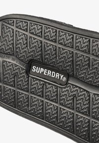 Zwarte rubberen zool met een gestructureerde grip, geometrische patronen en een gecentraliseerd zwart label met het "SUPERDRY"-logo in het wit.