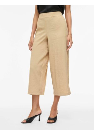 Pantalones beige de pierna ancha y corte corto combinados con mules negros de tacón y punta abierta, mostrados en una persona de pie sobre fondo blanco.