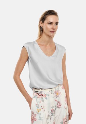 Donna che indossa un top senza maniche grigio chiaro con scollo a V infilato in pantaloni bianchi a vita alta con motivi floreali rosa e una cintura sottile.