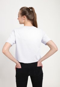 Biała krótka koszulka z okrągłym dekoltem, noszona z dopasowanymi czarnymi dżinsami. Modelka ma ręce w tylnej kieszeni, co podkreśla swobodny styl.