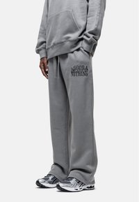 Grijze katoenen hoodie met een voorzak, gecombineerd met bijpassende grijze joggingbroeken met bedrukte tekst. Het schoeisel bestaat uit zwart en zilver hardloopschoenen.