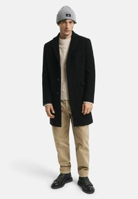 Schwarzer Wollmantel mit Reverskragen, beige gestrickter Pullover, khakifarbene Hose und schwarze Lederstiefel. Graue gestrickte Mütze auf dem Kopf.