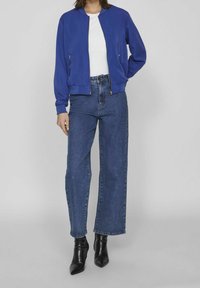 Chaqueta bomber azul con puños acanalados, combinada con unos jeans de mezclilla de talle alto y pernera ancha. El conjunto se completa con botas de tacón negro de punta.
