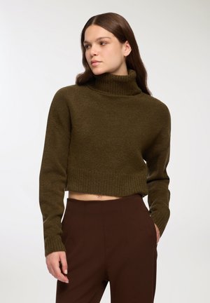 Donna che indossa un maglione corto a collo alto verde oliva e pantaloni marroni a vita alta, mentre guarda di lato con una mano in tasca.