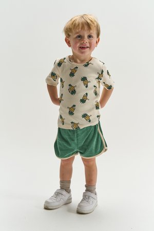 Fiatal fiú áll, bézs ingben, amelyen ananász minták vannak, zöld rövidnadrág, bézs zokni és fehér sportcipő visel, egyszerű háttér előtt.