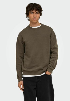 Jeune homme aux cheveux bouclés et foncés, portant un sweat marron et un pantalon noir, debout avec une main dans la poche devant un fond uni.