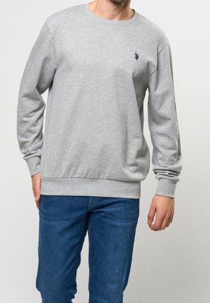 Hombre vestido con sudadera gris claro con un pequeño logotipo oscuro en el pecho y jeans azules, de pie contra un fondo liso.