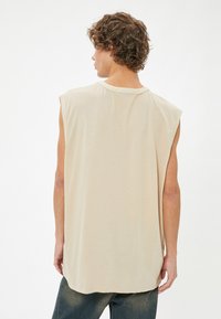 Beige mouwloze shirt met een ronde halslijn, zachte textuur en een iets langere achterkant, getoond vanuit de achterkant.