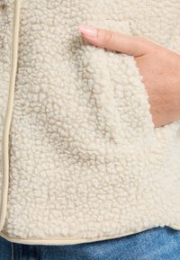 Beige fleece jacket with a textured surface, featuring a side pocket and contrast edging. Material appears soft and warm.