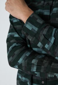 Vihreä ja musta ruudullinen paita, jossa on pienet mustat napit, pehmeä rakenne ja pitkät hihat, jotka osittain peittävät kyynärvarren.