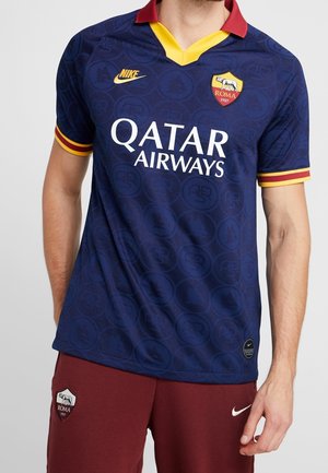 Mężczyzna ubrany w niebieską koszulkę piłkarską AS Roma z żółtymi i czerwonymi detalami oraz sponsorem Qatar Airways, zestawioną z bordowymi spodenkami AS Roma.