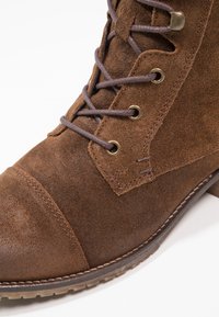 Hnědé semišové kotníkové boty s kovovými očky, hnědými tkaničkami, mírně zaoblenou špičkou a kontrastním prošíváním podél špičky a podrážky.