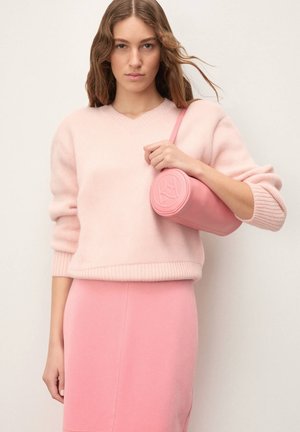 Světle růžový pletený svetr s výstřihem do V, sladěný s růžovou pouzdrovou sukní. Drží kulatou růžovou koženou kabelku s vypouklým logem.