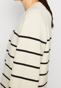 Suéter color crema con rayas horizontales negras, cuello redondo y puños acanalados. Textura suave con un ajuste relajado.