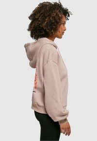 Light pink hoodie featuring a front pocket, drawstring hood, and side graphic design. Material appears soft with a relaxed fit.