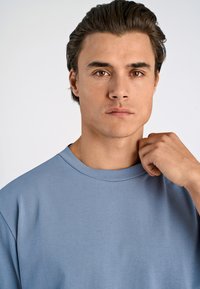 Jasnoniebieska bawełniana koszulka z okrągłym dekoltem i krótkimi rękawami, prezentowana na modelce, która lekko trzyma kołnierzyk jedną ręką.