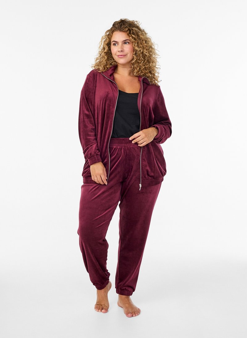 Woman with curly hair wearing a burgundy velour zip-up jacket and matching pants over a black top, standing barefoot on a white background.
