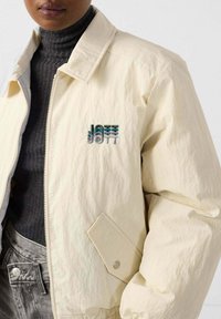 Giacca bomber beige chiaro con chiusura a zip, tasche laterali, colletto a coste e logo ricamato in blu e grigio sul petto.
