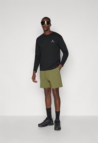 Man wearing black long-sleeve shirt, olive green shorts, black high socks, black athletic shoes, and large black sunglasses standing against white wall.