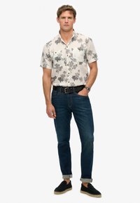 Camisa blanca de flores de manga corta con patrones en negro, combinada con unos pantalones vaqueros de color azul oscuro. Lleva un cinturón negro y zapatos negros tipo slip-on.