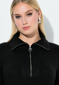 Zwarte geribbelde trui met een opstaande kraag en rits. Voorzien van een zilveren rits en een getextureerd breipatroon. Model heeft lang blond haar en zilveren oorbellen.