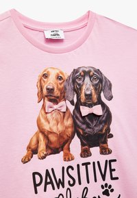 Camiseta rosa claro con un gráfico de dos perros que llevan corbatines rosas, uno de color marrón dorado y otro negro, con el texto "VIBRAS PAWSITIVAS."